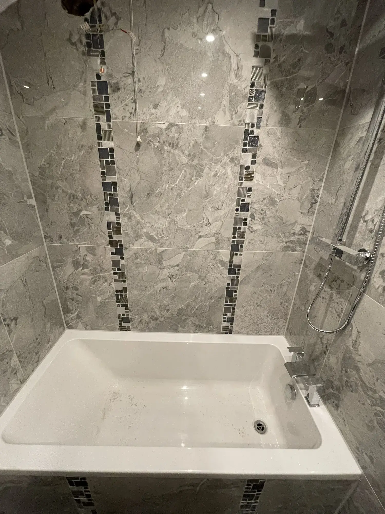 A newly installed white bath with a chrome bath filler against grey marble effect tiling.